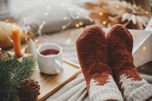 Piedi di donna in calzini di lana su un letto morbido con tazza di tè caldo, stelle di Natale, luci dorate, alberi, candele e cuscini. Accoglienti momenti invernali a casa. Ragazza che si rilassa nella stanza di sera scandinava