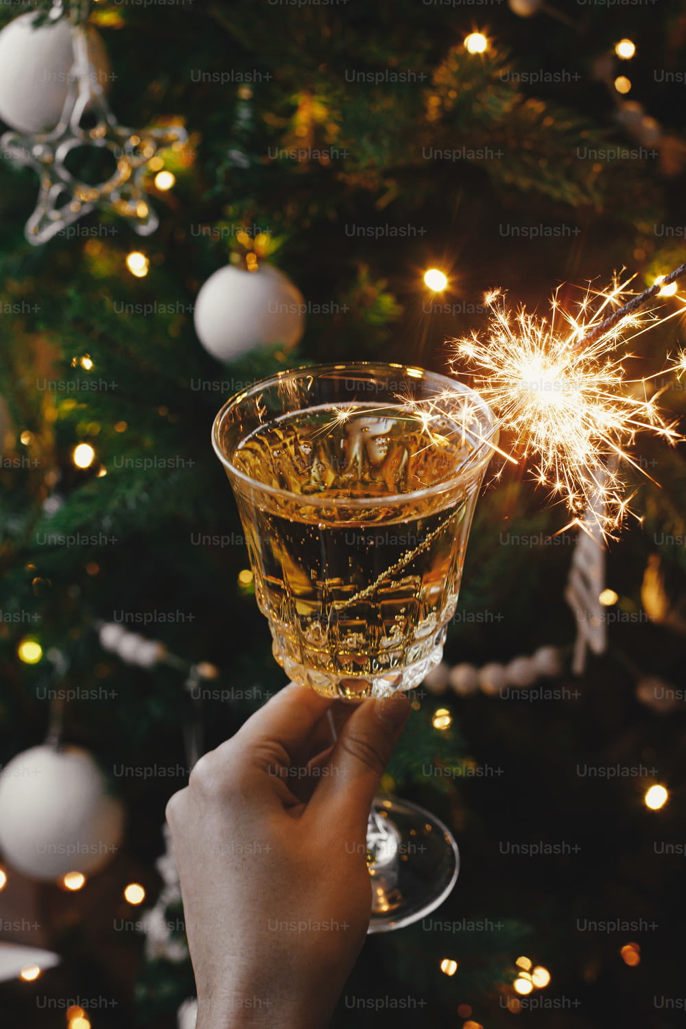Hand holding champagne glass with burning sparkler on background of christmas tree lights. Happy New Year! Firework bengal light and drink in woman hand. Atmospheric moment. New Year eve party