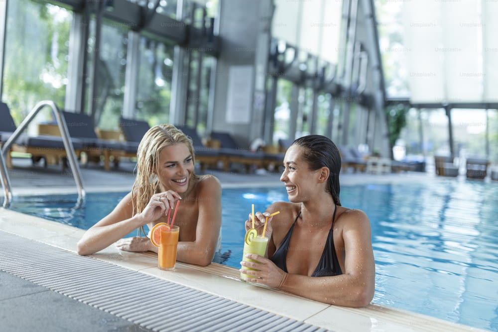 Poolside friends relaxing having healthy drinks. Sensual young women relaxing in spa swimming pool, spa indoor pool