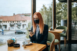레스토랑이나 카페 바에 앉아 스마트폰을 사용하는 젊은 우아한 비즈니스 여성. 그녀는 바이러스 질병에 대한 보호용 안면 마스크를 착용하고 있습니다. 코로나 바이러스와 새로운 정상 개념.