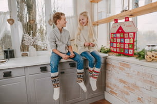 Garçon et fille heureux s’amusant assis sur la cuisine près du calendrier de l’avent fait à la main de Noël en forme de maison.