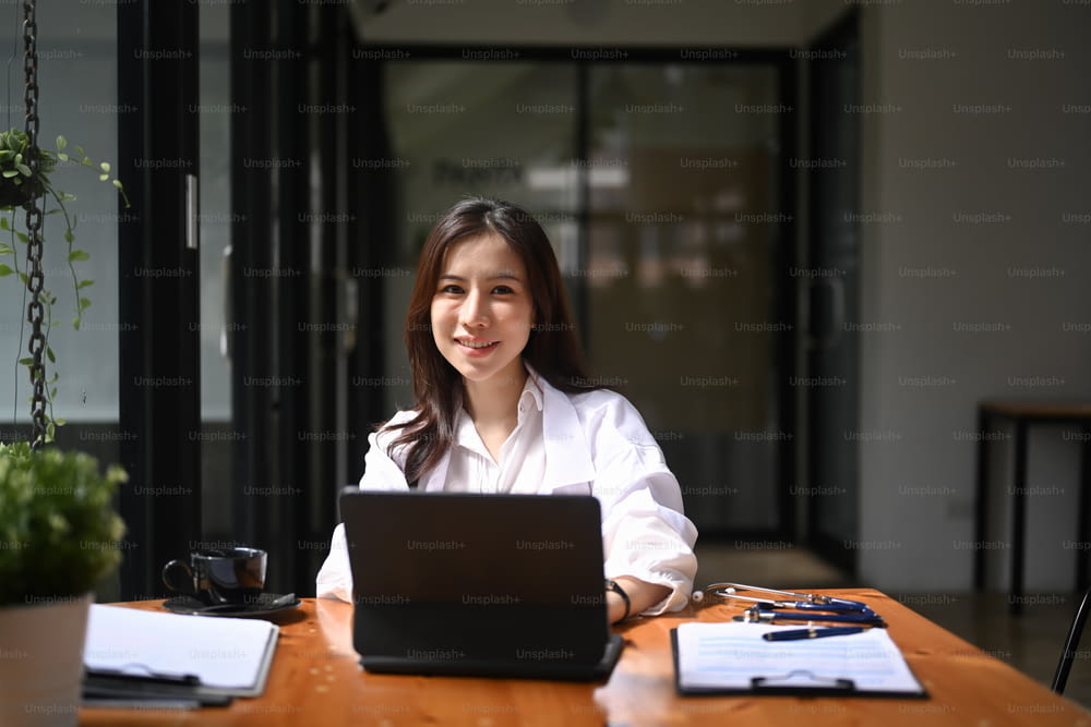 Femme médecin positive en uniforme médical blanc assise sur son lieu de travail et souriant à la caméra.