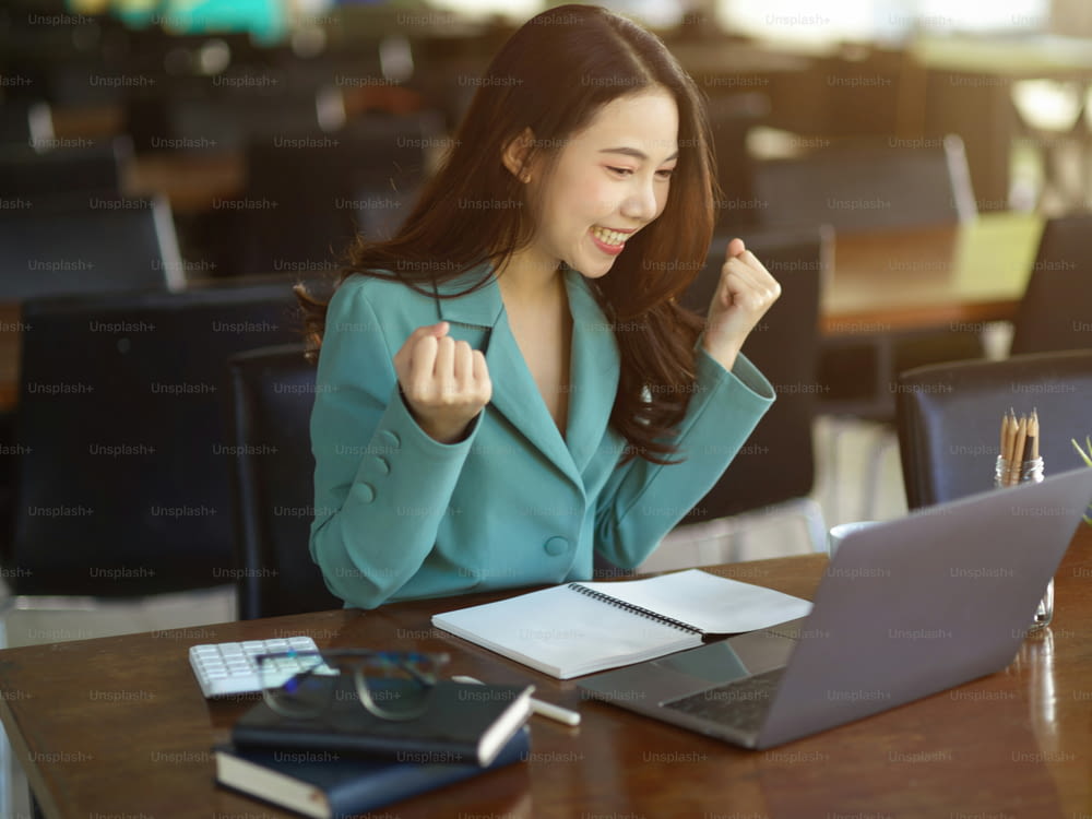 Successful business woman, female employee celebrating with raised hand while looking at laptop computer. Excited, Extremely happy.