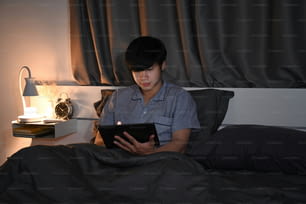Jeune homme en pyjama utilisant une tablette numérique sur son lit.