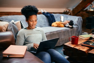 Mãe afro-americana navegando na internet no laptop enquanto sua filha está relaxando no sofá atrás dela.