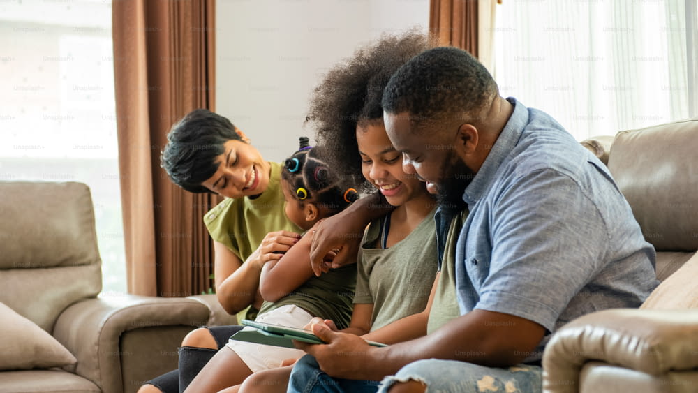 Pais mestiços africanos e duas filhas pequenas sentadas no sofá da sala de estar usando o tablet digital jogam ou assistem a filmes juntos. Família feliz desfruta da atividade de fim de semana com a tecnologia juntos em casa