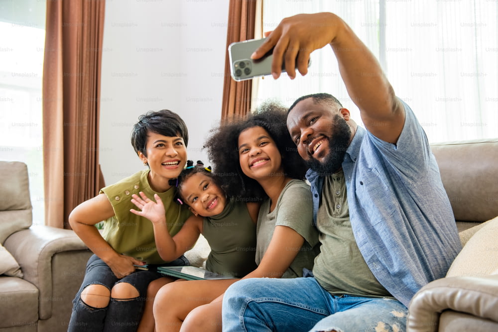 幸せなアフリカの両親と2人の小さな娘がリビングルームのソファに座って携帯電話を使って一緒に自撮りをしています。幸せな家族は、自宅でテクノロジーを使って楽しいレジャー活動を楽しんでいます
