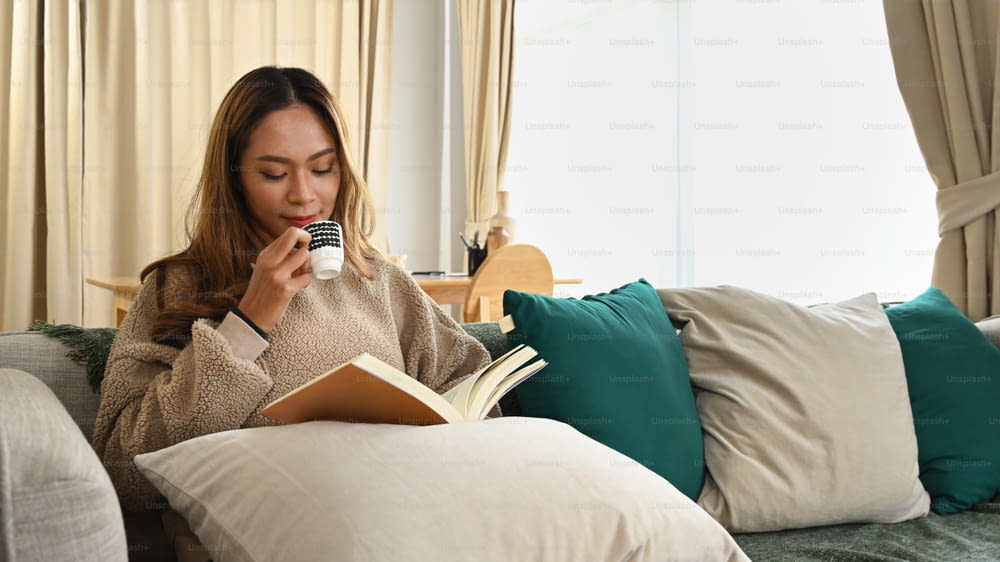 Peaceful asian woman reading book and drinking coffee on sofa.