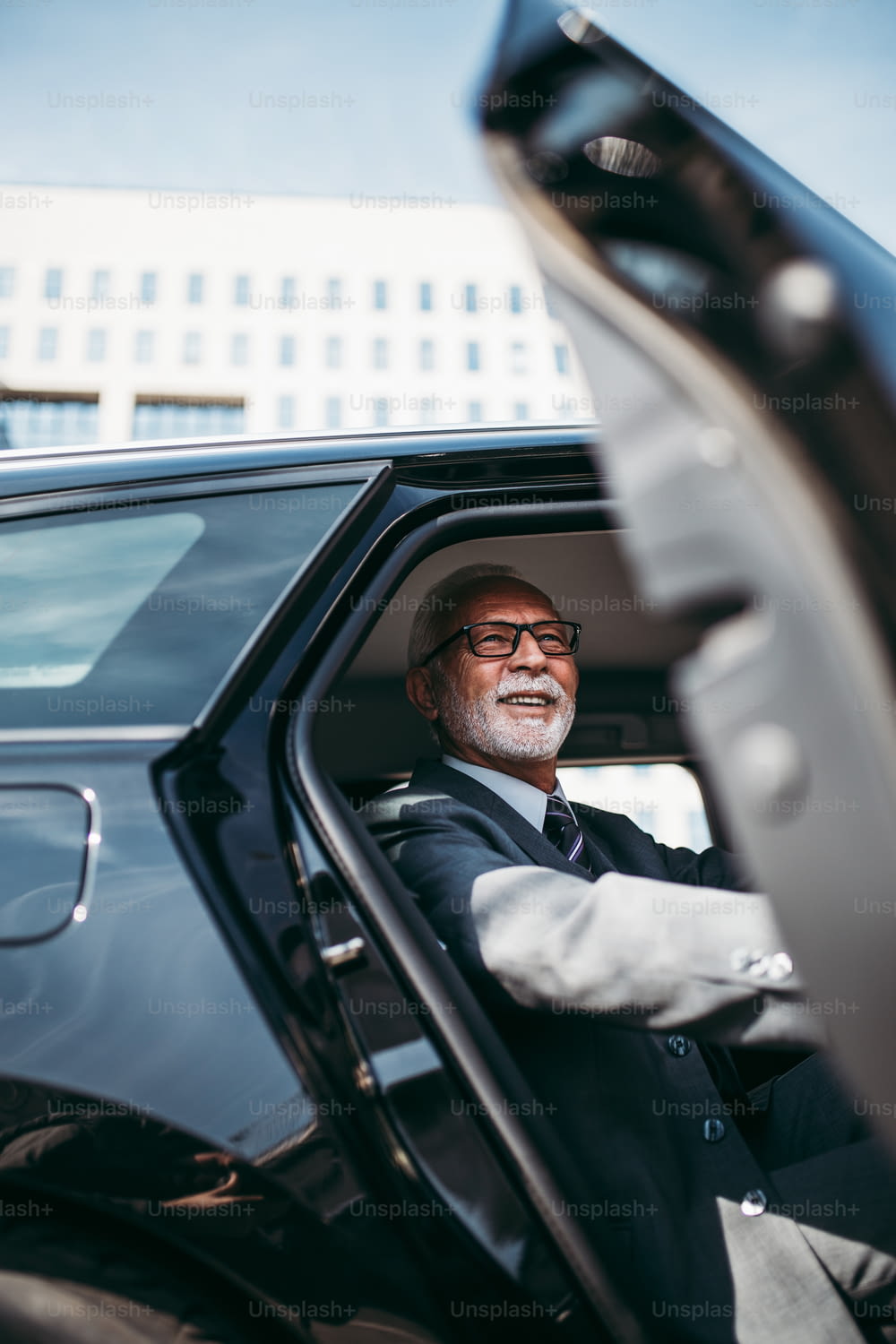 Good looking senior business man sitting on backseat in luxury car. He opens car doors and going or stepping out. Big busness bulding in background. Transportation in corporate business concept.