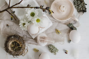 Natural easter eggs, feathers, willow branches, nest and candle on rustic white table flat lay. Stylish rural Easter still life in pastel white and beige colors. Modern simple aesthetics