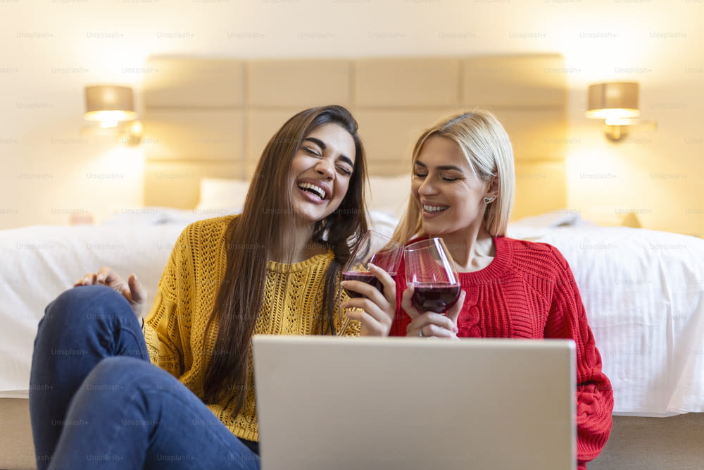 Two attractive girls, cheerful best friends having fun,watching movies on laptop and drinking red wine at home. Two glasses of white wine in hands