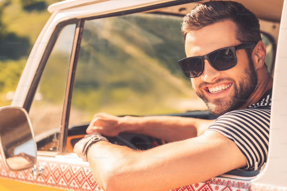 Jovem alegre sorrindo para a câmera e segurando a mão no volante enquanto está sentado dentro de sua minivan