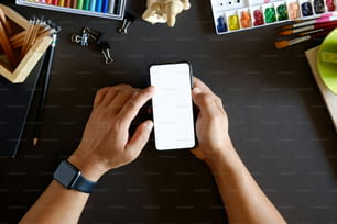 Cropped and top view shot of man using a smartphone over dark designer desk,  laptop, artist craft supplies ,Mobile phone   has blank mockup screen.