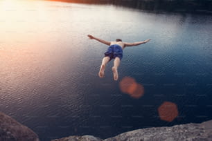 Junger Mann springt bei Sonnenuntergang mit ausgebreiteten Händen von der Klippe ins Wasser (absichtliche Sonnenblendung und Lensflares, Mann in Bewegungsunschärfe)