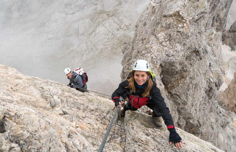 young attractiv climbers on a vertical and exposed rock face climbs a Via Ferrata while smiling and pointing to a faraway mountain peak