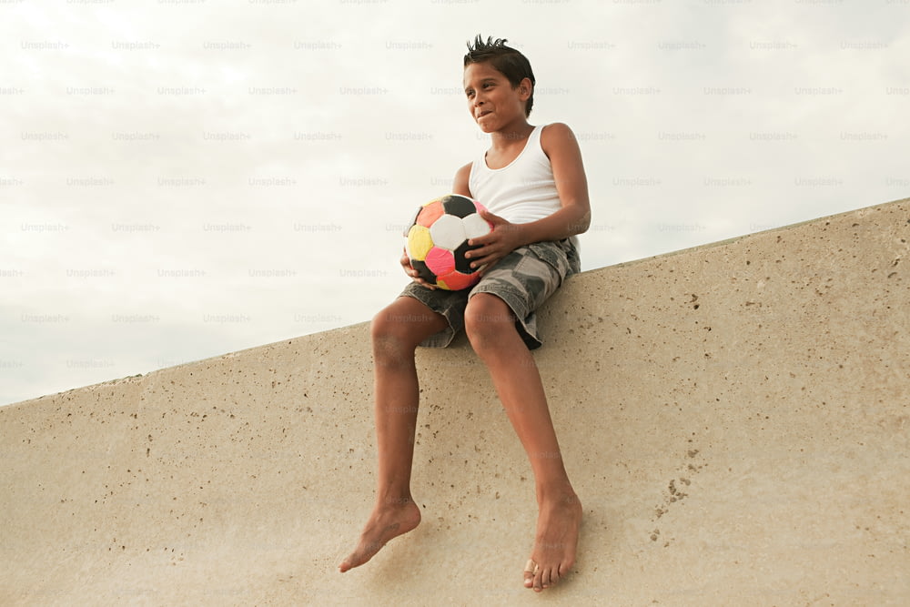 un ragazzo seduto su un muro che tiene un pallone da calcio