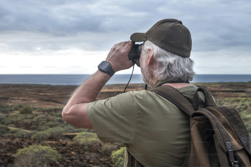 Rear view of senior man with white beard looking at the horizon with binoculars. Active retiree with backpack on shoulders enjoying the hike