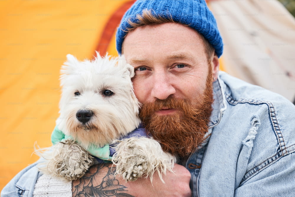 Look at us! Bearded man looking to the camera while cuddling his white terrier dog. They are best friends. Stock photo