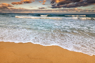 Vista grandangolare di un'idilliaca spiaggia sabbiosa con un'onda azzurra in arrivo con l'alta marea. Tempo ventoso e tempestoso al tramonto. Sfondo per viaggi e vacanze