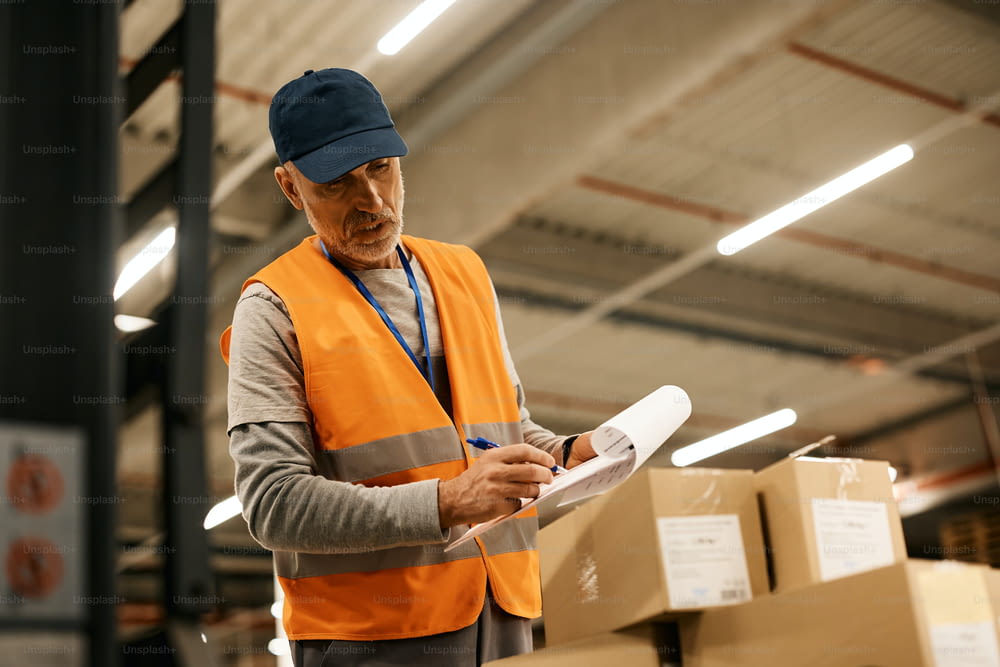 Mature worker writing on clipboard while preparing packages for distribution while working in a warehouse.