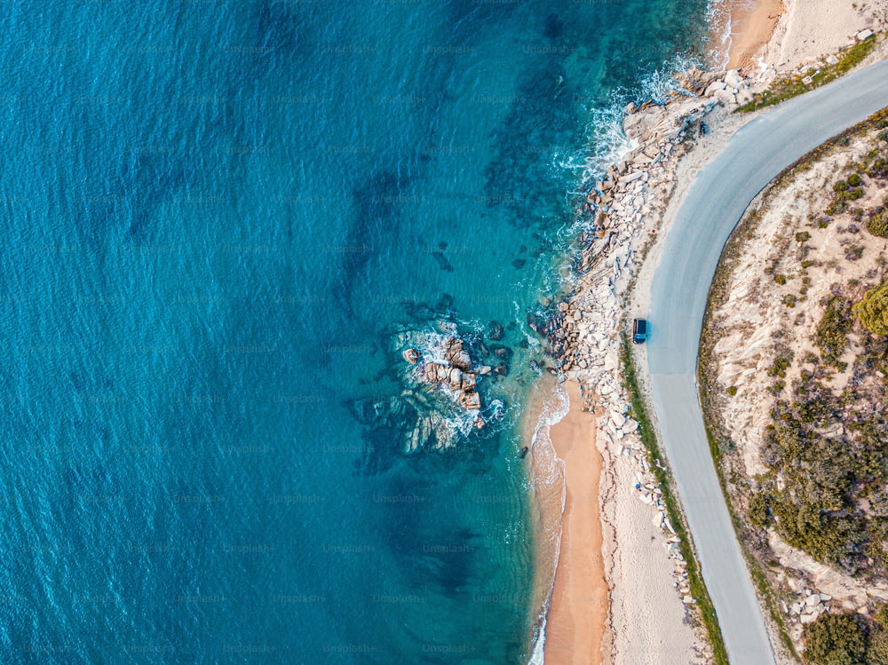 Luftaufnahme eines idyllischen Meeressandstrandes mit asphaltierter kurvenreicher Straße und weiterfahrendem Kleinwagen. Hintergrund für Reisen und Urlaub