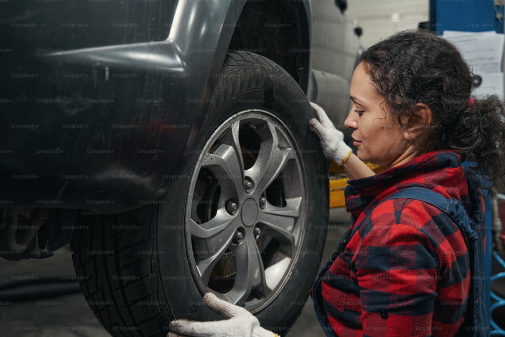 Female vehicle technician in work gloves inspecting automobile wheel in car garage