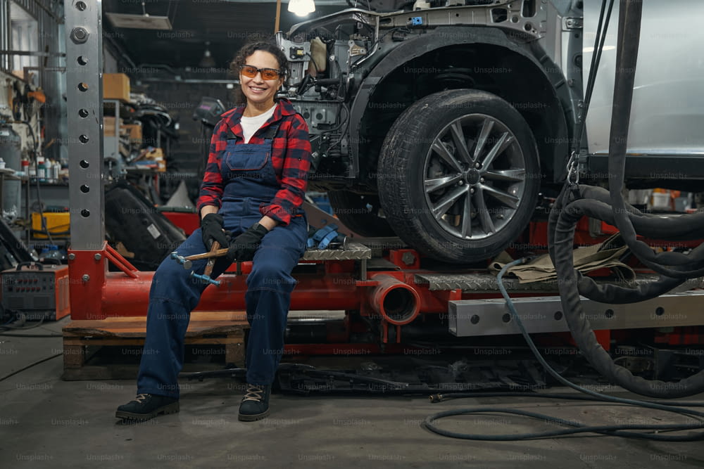 Female worker in safety glasses holding hammers and smiling while resting near automobile in garage