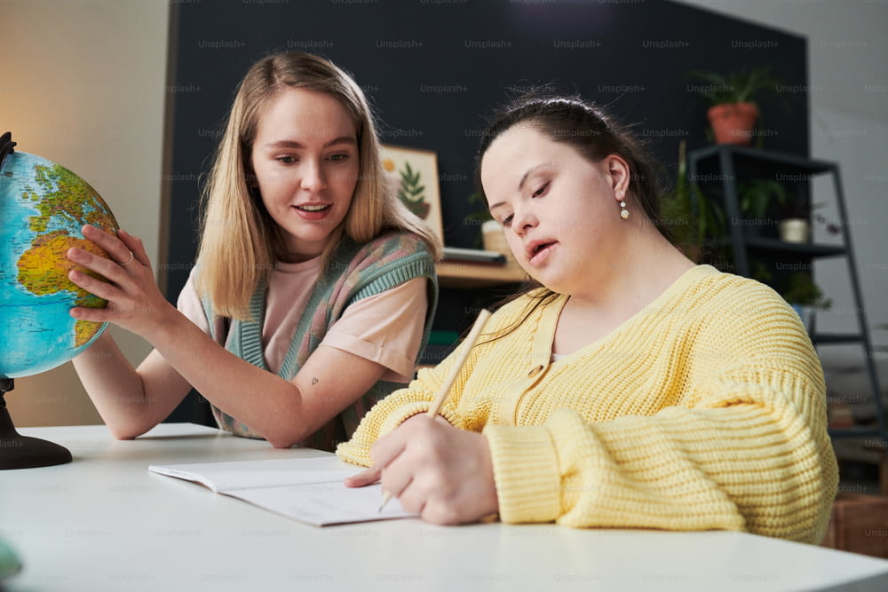 Young female teacher sitting at table next to her student with Down syndrome helping with task during geography lesson