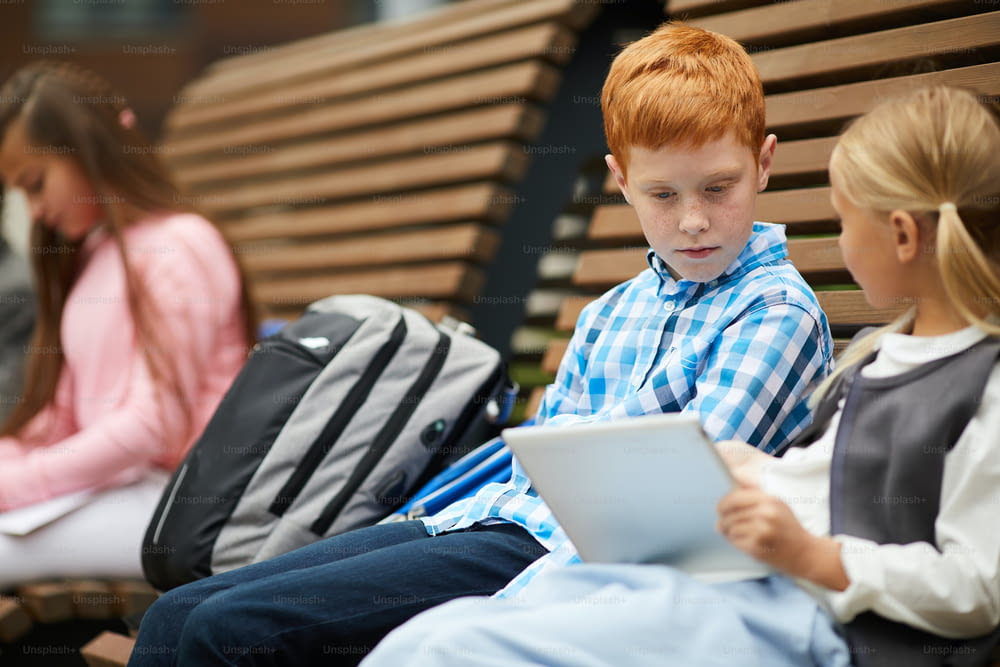 Studentessa con tablet pc che mostra qualcosa allo scolaro dai capelli rossi mentre si siedono insieme sulla panchina all'aperto