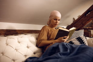 Woman suffering from cancer reading book while relaxing on the sofa at home. Copy space.