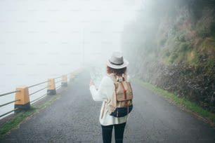 Ragazza viaggiatrice riccia che indossa zaino e cappello in piedi sulla strada nella nebbia, cercando il sentiero e usando la mappa.