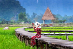 Junge Frau sitzt auf Holzweg und fotografiert mit grünem Reisfeld in Vang Vieng, Laos.