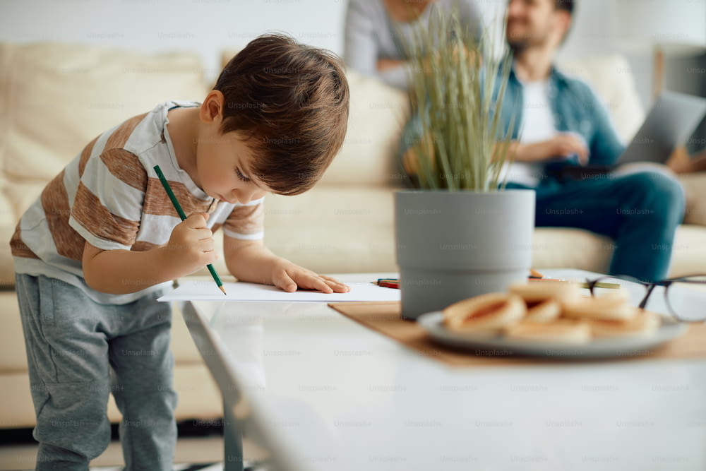 Création d’un petit garçon qui fait des croquis sur une feuille de papier à la maison. Ses parents sont à l’arrière-plan.