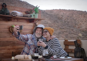 Two elderly people in love look at the cellphone for a selfie. Food and drink on the table with salami and red wine. Mountain in background