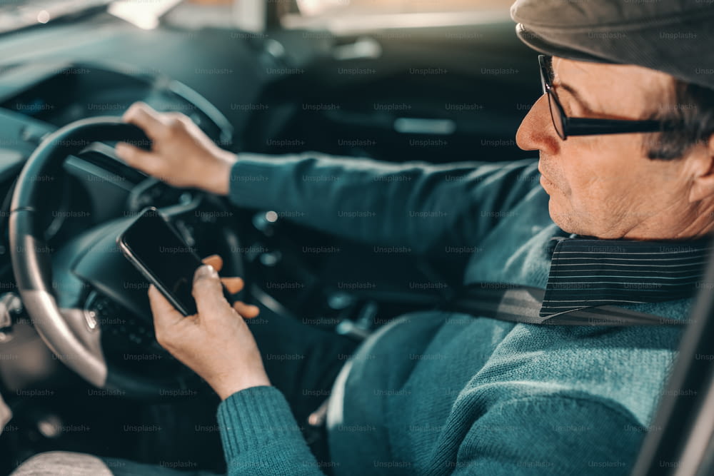 Caucasian senior with hat on head and eyeglasses driving car and using smart phone.