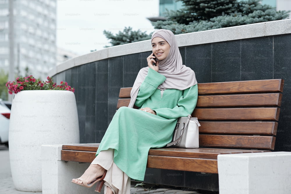 Smiling stylish young Muslim businesswoman in long green dress and hijab sitting on bench with bag and calling by phone