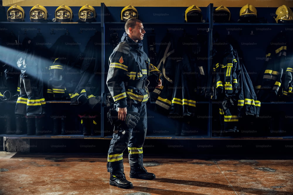 Giovane vigile del fuoco coraggioso in piedi nella caserma dei vigili del fuoco in uniforme protettiva e tenendo il casco sotto l'ascella.