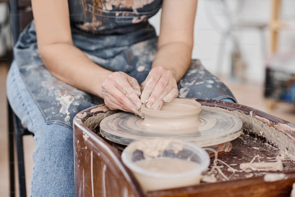 Gros plan d’une jeune artisane créative habile à la poterie créant un nouvel objet en argile tout en étant assise près de la roue rotative dans l’atelier