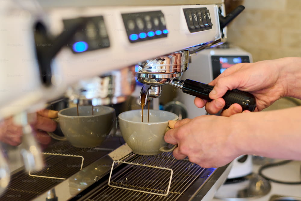 カップを持ち、カフェのイナゴマメのコーヒーマシンでコーヒーを準備するプロのバリスタの接写