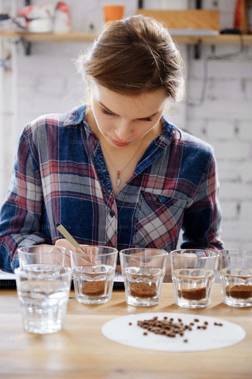 Portrait of young female barista writing result of coffee cupping test, examining cups with beans on wooden table