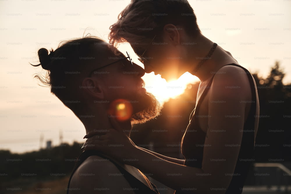 夕焼けの自然光の中で屋外でキスをするカラフルな髪を持つ丸い眼鏡をかけた若い夫婦の接写。太陽の光がそれらの間を照らします