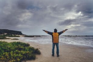 Travel explore destination with one man viewed frmo back outstretching arms for satisfaction and happiness in fron tof a wild bad weather beach and dramatic sky with ocean