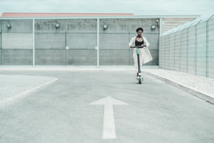Una joven africana elegante con gafas y una capa blanca ondeante conduce un e-scooter en la carretera de asfalto con una flecha pintada como marca de carretera en un primer plano desenfocado, poca profundidad de campo