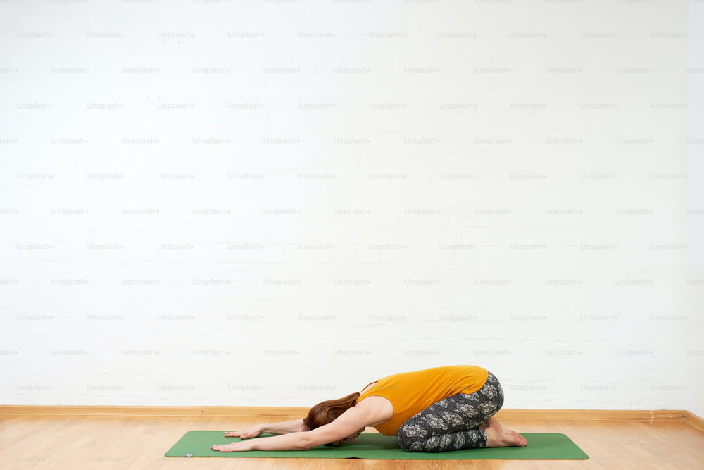 Imagen de cuerpo entero de una mujer de mediana edad haciendo yoga flow en una estera verde cerca de la pared blanca, lleva un tanque amarillo y pantalones oscuros