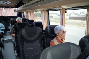 Aged Caucasian woman with grey hair sitting by window inside bus and looking at road and country houses