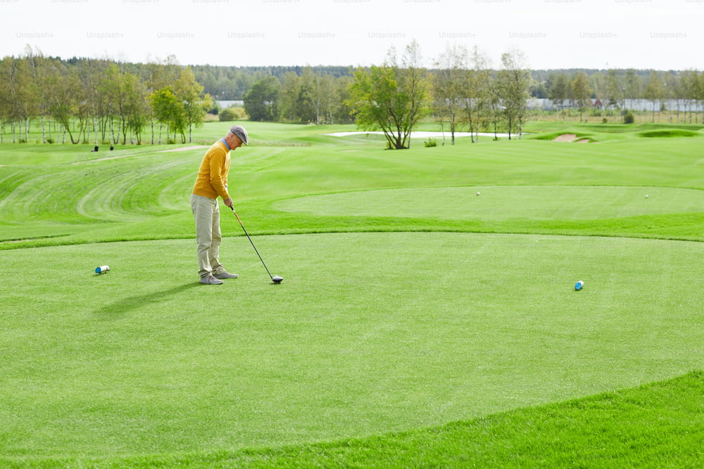 Mature man with golf club going to hit ball while standing on green lawn on play area