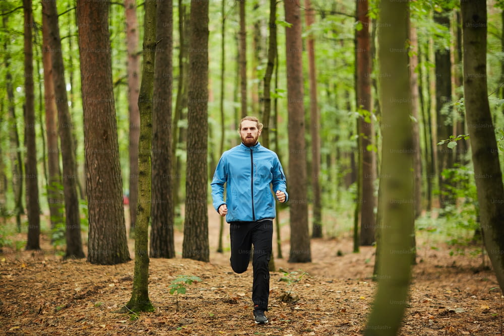 Young sportsman running among pines and birch trees along forest path while training in the morning