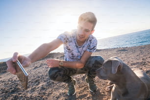가장 친한 친구 함께 젊은 백인 남자와 회색 아름다운 amstal 개는 배경에 햇빛이 비치는 일몰이 있는 야외에서 스마트폰으로 셀카 사진을 찍고 있다 - 바다와 휴가 개념