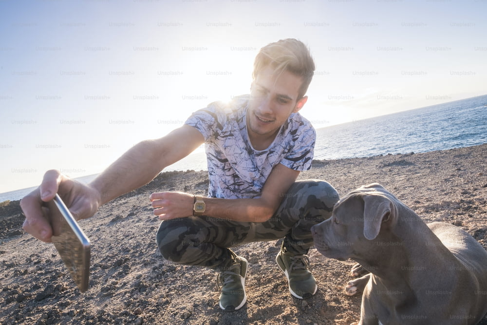 Melhores amigos juntos jovem homem caucasiano e cinzento belo cão amstal tirando selfie foto com telefone inteligente ao ar livre com pôr do sol luz do sol no fundo - oceano e conceito de férias