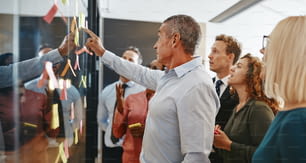 성숙한 관리자와 그의 팀은 현대적인 사무실에서 함께 일하면서 유리 벽에 스티커 메모로 브레인스토밍합니다.